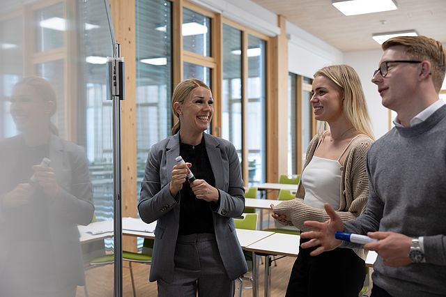 Lernsituation: Eine Dozentin steht mit zwei ihrer Studierenden vor einem Whiteboard, um Lerninhalte zu besprechen.