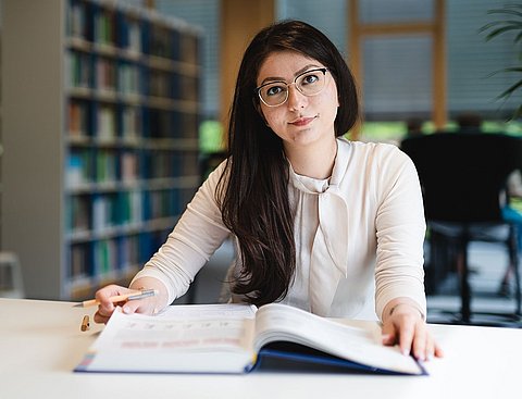 Eine junge Frau sitzt in einer Bücherei an einem Tisch mit einem Buch vor sich.