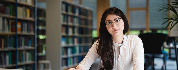 Eine junge Frau sitzt in einer Bücherei an einem Tisch mit einem Buch vor sich.