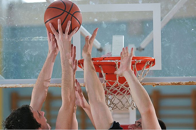 Im Fokus des Bildes sind die Arme mehrerer Spieler, die versuchen, den Basketball in den Korb zu manövrieren, oder die versuchen, ihn zu blocken.