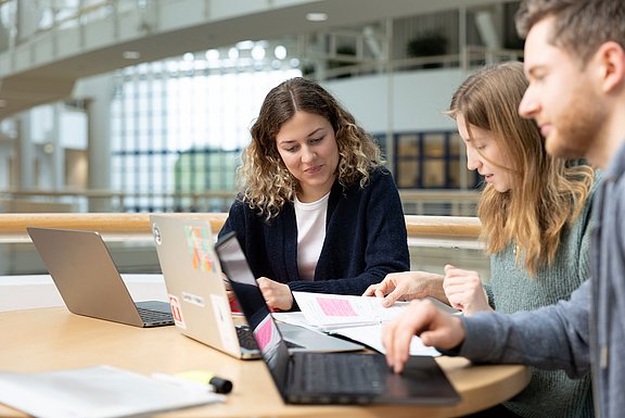 Drei junge Menschen sitzen an einem Tisch und schauen gemeinsam auf ihre Laptops.