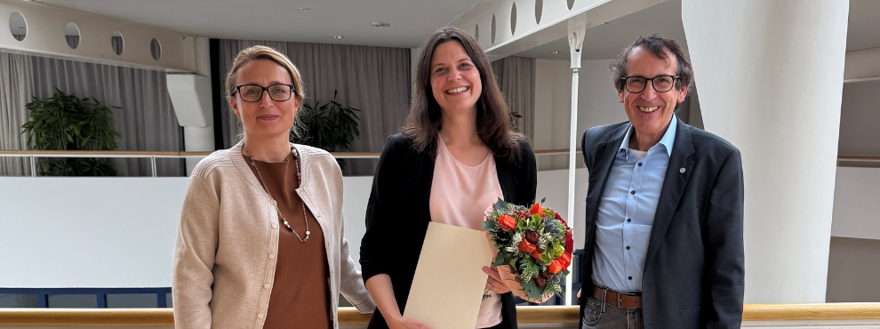 Drei Personen stehen im Hauptgebäude der Uni Witten/Herdecke. Die Person in der Mitte ist Prof. Dr. Anke Haberkamp. Sie hält ihre Berufungsurkunde in der Hand und lächelt freundlich in die Kamera.