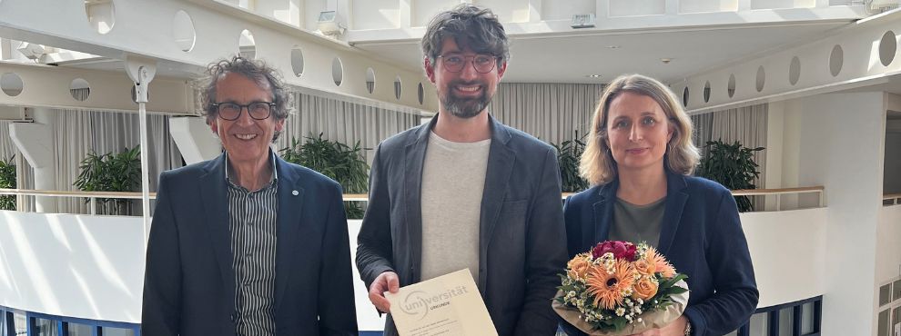 Three people smile frontally into the camera. The man in the middle holds a certificate into the picture.