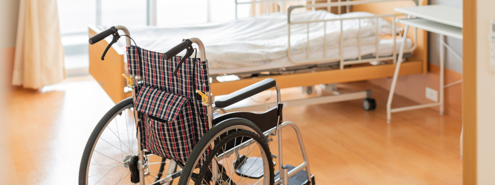 A room in a care home. A wheelchair stands in front of an empty bed.