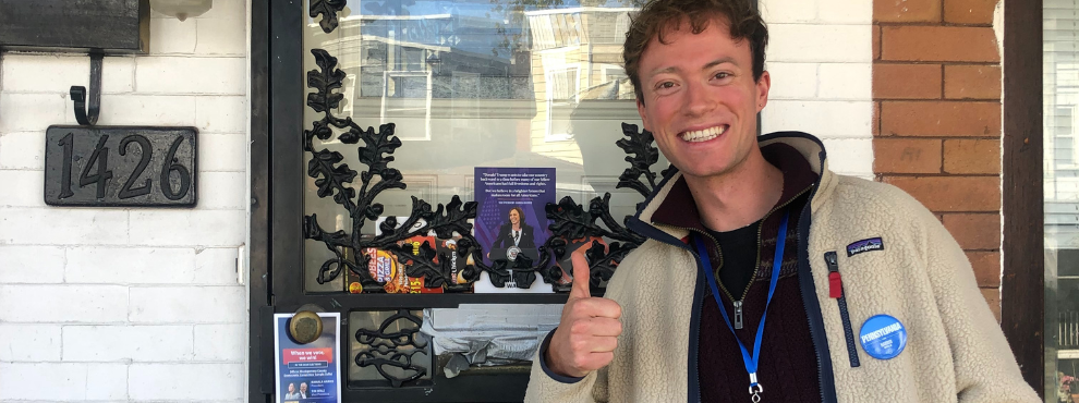 A young man stands in front of a front door with his thumb up and laughs into the camera. He is wearing a blue election worker button. Next to him on the door are election flyers for presidential candidate Kamala Harris.