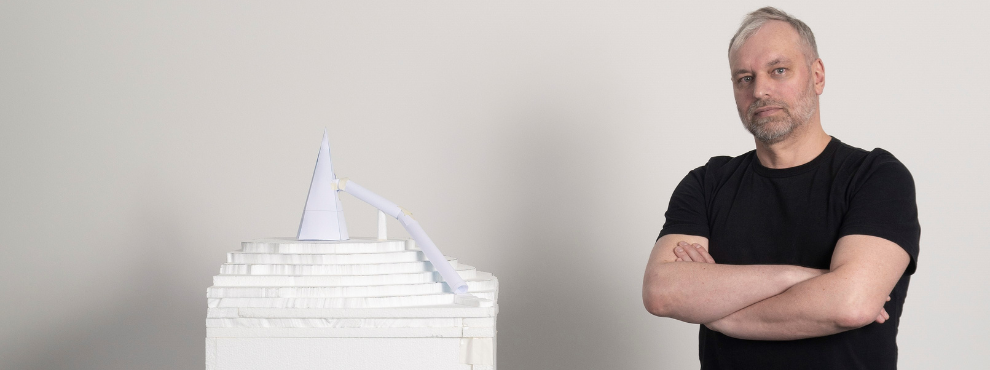 A man with a black T-shirt and folded arms sits next to a white model of a slide that comes out of a 3D printer.