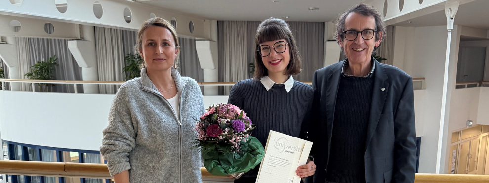 Drei Personen stehen nebeneinander in der Universität. In der Mitte Theresa Busse, eine junge Frau, die ihre Berufungsurkunde und einen Blumenstrauß in der Hand hält und glücklich aussieht. Links von ihr Prof. Dr. Margareta Halek, rechts Prof. Dr. Stefan Zimmermann.